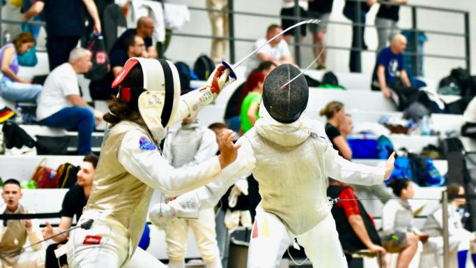 Zwei Fechter in Wettkampf, umgeben von Zuschauern und Sportlern.