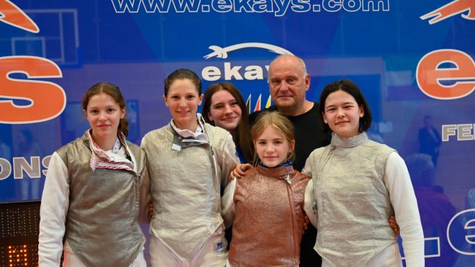 Gruppenfoto von fünf Fechterinnen und einem Trainer, lächelnd vor einem blauen Hintergrund.