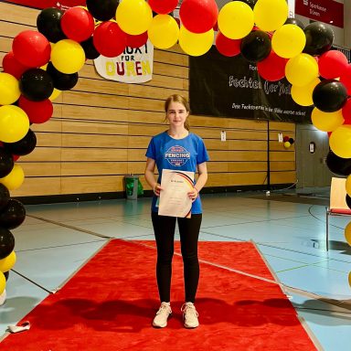 Junge Frau steht unter einem bunten Bogen aus Luftballons auf einem roten Teppich.