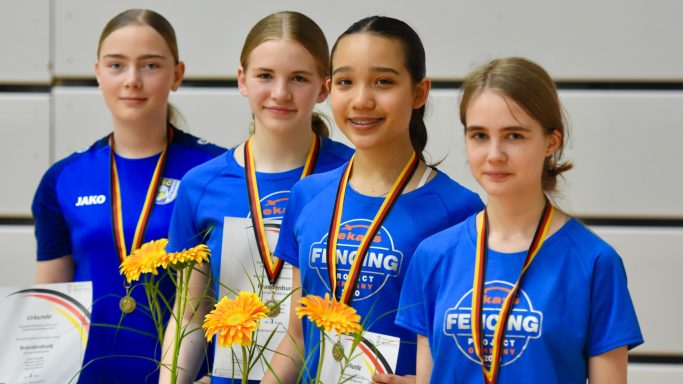 Vier junge Sportlerinnen mit Medaillen und Urkunden, lächelnd in blauen T-Shirts.